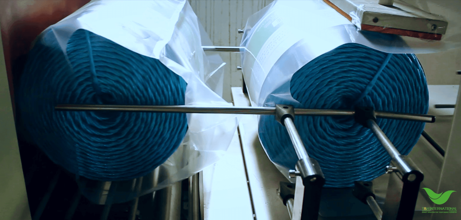 Two large rolls of blue industrial material on metal rods, partially wrapped in clear plastic.